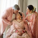 Bride's father kissing on her forehead and mother standing next tot her, all in light pink dress in front of window.
